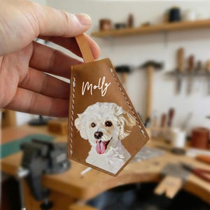 May include: A brown leather keychain with a painted portrait of a white dog with its tongue out. The name "Molly" is written in white script above the dog. The keychain has a leather loop.