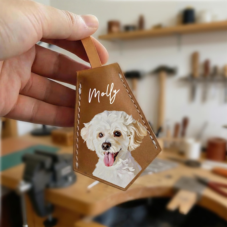 May include: A brown leather keychain with a painted portrait of a white dog with its tongue out. The name "Molly" is written in white script above the dog. The keychain has a leather loop.