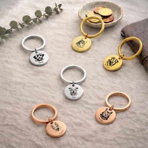 May include: A collection of personalized pet keychains in gold, silver, and rose gold. Each keychain features a circular pendant with a detailed engraved portrait of a dog and the pet's name. The keychains are arranged on a neutral surface.