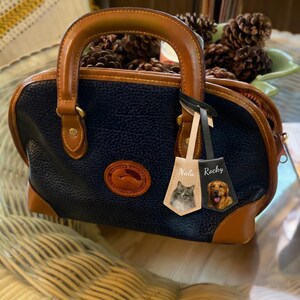 May include: A navy blue and brown leather handbag with a rounded shape and a top handle. The bag features a logo patch and two personalized tags, one with a cat and the name "Nala", and the other with a dog and the name "Rocky".
