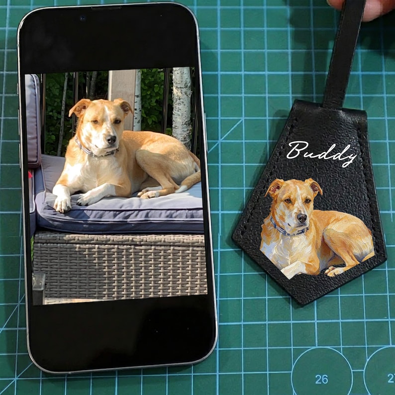 May include: A tan and white dog is captured in a photo on a smartphone screen, and also on a black leather tag. The tag has the dog's image and the name "Buddy" in white script. The dog is wearing a blue collar.