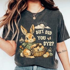 Puede incluir: Camiseta gris oscuro con un diseño de Pascua de estilo vintage. El gráfico incluye un conejito sosteniendo una cesta de huevos decorados, con el texto "BUT DID YOU DYE?" encima del conejito. Un sol y nubes también están presentes.