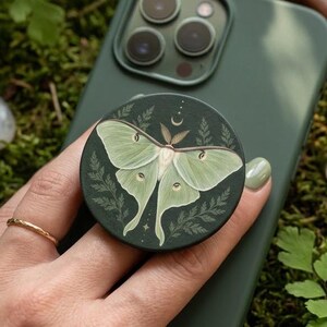 May include: A circular phone grip featuring a detailed illustration of a pale green luna moth centered on a dark green background, accented with delicate fern leaves, a small crescent moon, and tiny stars above the moth's wings.