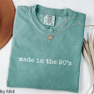 Puede incluir: Una camiseta verde agua doblada con la frase "made in the 90's" en letras blancas. Un collar plateado con un pequeño colgante descansa sobre la camiseta. La camiseta tiene una etiqueta "Comfort Colors".