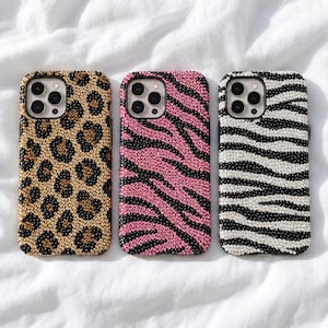 May include: Three smartphone cases featuring a camera cutout and a surface covered in small, reflective beads. The left case has a gold and black leopard print, the middle case has a pink and black tiger stripe pattern, and the right case has a white and black zebra stripe pattern.