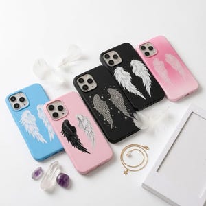 May include: Five smartphone cases in blue, pink, black, and light pink, each featuring angel wing designs. One black case displays rhinestone-encrusted wings, while others show printed white or black wings. White feathers and crystals are scattered nearby.