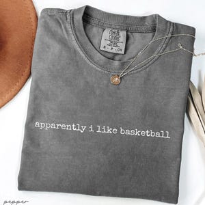 May include: A gray Comfort Colors t-shirt with the phrase "apparently i like basketball" in white lettering. A silver necklace with a circular pendant rests on the shirt. A brown hat is visible in the upper left corner.