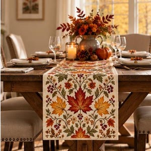 May include: A rectangular table runner featuring a cream-colored background with a repeating pattern of vibrant red, orange, and yellow maple leaves, green foliage, and small clusters of red berries, draped over a wooden dining table.