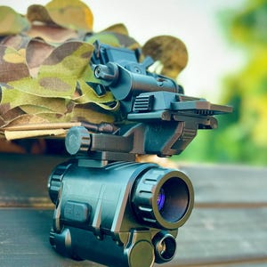 May include: A black night vision device with a camouflage cover. The device has a large lens and several adjustment knobs. The background is blurred, with green foliage visible.
