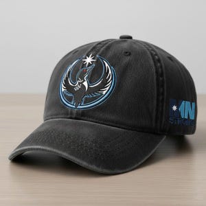 May include: A charcoal gray baseball cap featuring a circular emblem with a bird design in black, white, and light blue. The text "MN STRONG" is embroidered on the side in blue. The cap has a curved brim and a button on top.