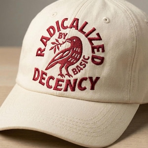 May include: Cream-colored baseball cap with the embroidered text "RADICALIZED BY BASIC DECENCY" in burgundy. A burgundy bird holding a sprig is centered on the cap.