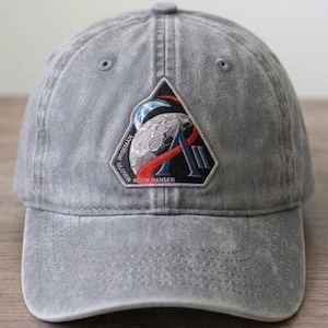 May include: Gray baseball cap with a patch featuring a stylized moon, Earth, and the text "Artemis III." The patch also includes the names Wiseman, Glover, Koch, and Hansen. The cap has a curved brim and adjustable strap.
