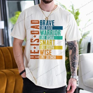 May include: Cream-colored t-shirt with the text "HE IS DAD" in a vertical orange font, and the words "BRAVE, WARRIOR, SMART, WISE" in a stacked design. Each word is followed by "LIKE" and a name, in a different color.