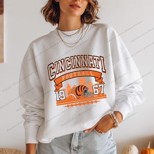 May include: White sweatshirt featuring a Cincinnati Football design in orange and black. The graphic includes the city skyline, a football helmet, and the year 1967. The sweatshirt has long sleeves and a crew neck.