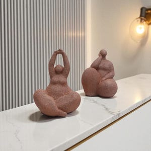 May include: Two terracotta-colored abstract sculptures of seated figures. One figure has arms raised overhead, the other rests a hand on its cheek. The sculptures are on a white surface, with a striped wall in the background.