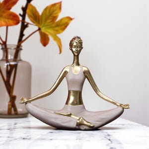 May include: A gold and beige yoga pose figurine. The figurine is in a seated lotus position with arms outstretched. The figure's top and waist are gold, while the rest of the figure is beige. A vase with fall leaves is in the background.