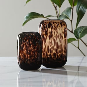 Puede incluir: Dos jarrones decorativos de cristal. Los jarrones son cilíndricos con bordes redondeados y un patrón moteado en tonos marrones y ámbar. El jarrón más pequeño es marrón oscuro y el más grande es ámbar. Los jarrones se exhiben sobre una superficie blanca.