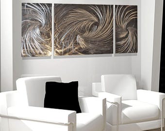 May include: A three-piece metal wall art set featuring a swirling abstract design in shades of silver and gold. The artwork is displayed on a white wall above two white chairs with black throw pillows.