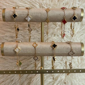 May include: Gold bracelets with clover-shaped pendants in black, white, red, and gold are displayed on a jewellery stand. The bracelets hang from a cream-coloured, cylindrical display.