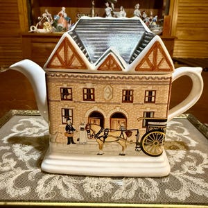 May include: A ceramic teapot shaped like a two-story building with a dark gray roof. The exterior features a tan brick facade with brown trim, windows, and a scene depicting a horse-drawn carriage with the text RINGTONS printed on the side.