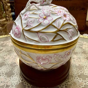 May include: A white porcelain lidded bowl with pink floral and gold leaf designs. The bowl sits on a dark wooden base. The lid has a small, decorative handle. The word "HERBS" is visible in the background.