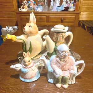 Può includere: Una collezione di quattro teiere in ceramica con disegni di personaggi. Una è un grande coniglio che tiene un fiore giallo, una è una persona con un cappello, un piccolo coniglio con un vestito blu e rosa e una persona anziana seduta su una sedia.