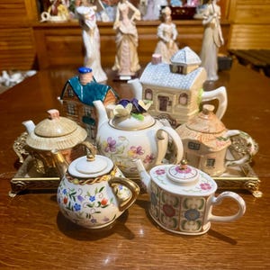 Peut inclure: Une collection de théières en céramique de formes variées, incluant des modèles en forme de maisonnettes et des motifs floraux, disposées sur un plateau rectangulaire en métal doré. Les théières présentent des détails peints, des accents dorés et des couvercles, avec des couleurs comme le bleu, le vert, le rose et le violet sur des fonds blancs et beiges.