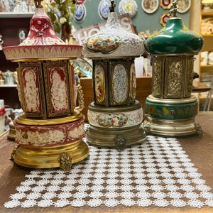 May include: Three antique music boxes with ornate designs. The boxes feature detailed relief carvings, painted scenes, and decorative metal accents. One has a red top, another a white top, and the third a green top. They sit on a wooden table with a white lace runner.