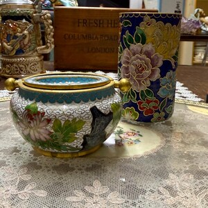 May include: An antique collection featuring a cloisonné bowl with floral designs, a blue and gold vase, and a wooden box with text. The bowl has a turquoise interior and gold accents. The vase displays colorful flowers. A magazine titled "Homes & Antiques" is visible.
