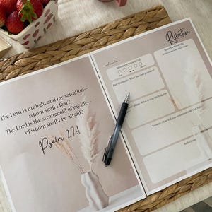 May include: Two open pages of a journal with the text "The Lord is my light and my salvation - whom shall I fear?" and "Psalm 27:1". A pen rests on the page. A small vase with dried flowers is on the left. A cup and a bowl of strawberries are in the background.