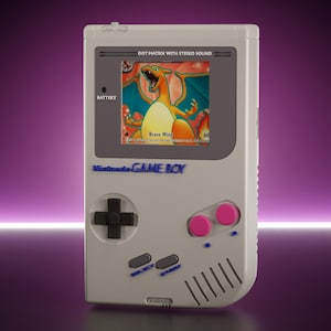 Puede incluir: Una consola de videojuegos portátil de color gris claro con un marco de pantalla gris oscuro, una cruceta negra, dos botones de acción rosas y dos botones select y start grises. La pantalla muestra un personaje Charizard con el texto Brave Wing.