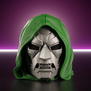 Op de afbeelding: Een zilverkleurig metalen masker met een stijf, hoekig ontwerp met zichtbare klinknagels en donkere oogopeningen, gedeeltelijk bedekt door een gestructureerde, felgroene kap die over de bovenkant en zijkanten van het gezichtsdeel valt.