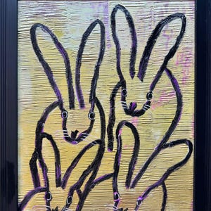 Puede incluir: Un cuadro enmarcado que muestra cuatro conejos delineados con trazos de pintura negra gruesa sobre un fondo texturizado dorado y amarillo. Los conejos tienen orejas largas y rasgos simples, con toques de pintura morada en los contornos.