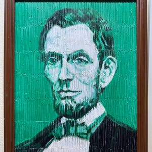 Puede incluir: Una pintura enmarcada que presenta un retrato de Abraham Lincoln realizado en tonos blancos y verde oscuro sobre un fondo verde vibrante. La obra muestra una técnica de pinceladas verticales texturizadas en toda la composición.