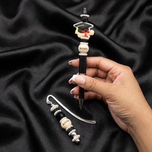 Pode incluir: Um conjunto de caneta e marcador de livros pretos com enfeites com tema de formatura. A caneta apresenta um design de chapéu de formatura e a palavra "BLESSED" em uma conta. O marcador de livros tem contas semelhantes e a palavra "BLESSED".