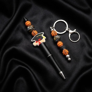 May include: A black pen and keychain set with basketball-themed beads. The pen features a graduation cap charm. The keychain has a silver ring and clasp. The beads are orange, black, and silver.