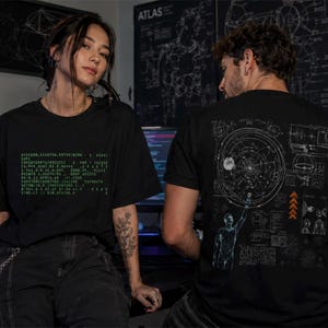 May include: Two people wearing black t-shirts. One shirt has green text, the other has a diagram on the back. The background includes computer monitors, a desk, and a wall with diagrams and text.