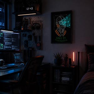 Peut inclure: Une affiche murale encadrée représentant une illustration de tête robotique stylisée avec des accents orange et bleus, étiquetée UNIT 01 ATLAS PROTOCOL. L'œuvre est exposée dans une pièce sombre avec un bureau, un panneau perforé et une étagère.