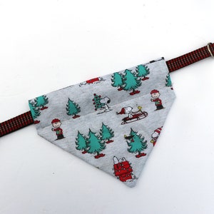 May include: Gray dog bandana with a Christmas-themed pattern featuring Snoopy, Charlie Brown, Christmas trees, and other Peanuts characters. The bandana is attached to a red and black striped collar with a black buckle.