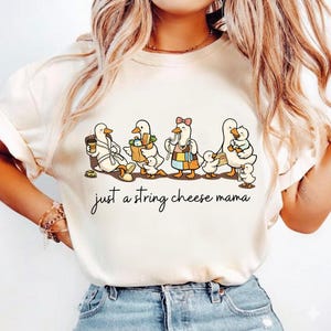 May include: Cream-colored t-shirt featuring a whimsical illustration of cartoon ducks in various outfits. The ducks are depicted with groceries, coffee, and a bow. The text "just a string cheese mama" is written in cursive below the ducks.