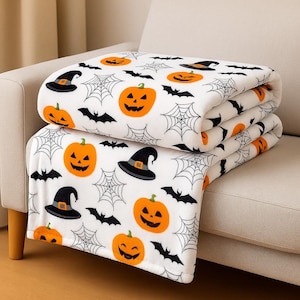 May include: A folded white Halloween-themed blanket featuring orange jack-o'-lanterns, black bats, witch hats, and spiderwebs. The blanket is made of a soft material and is folded neatly on a beige couch.