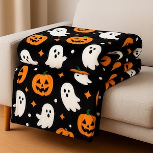 May include: A black Halloween blanket with a repeating pattern of white ghosts and orange pumpkins. The blanket is folded and placed on a beige sofa. The design includes orange stars and white dots.