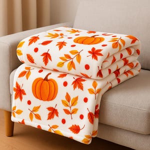 May include: A folded white blanket featuring a pattern of orange pumpkins, red dots, and autumn leaves in orange and yellow hues. The blanket is folded on a grey sofa, highlighting its seasonal design, perfect for autumn.