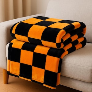 Peut inclure: Deux couvertures en peluche avec un motif à carreaux noir et orange sont pliées sur un canapé beige. Les couvertures ont une texture douce et veloutée et sont probablement faites d'un matériau chaud. Le schéma de couleurs est audacieux et accrocheur.