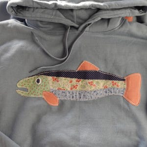 Peut inclure: Un sweat à capuche bleu clair orné d'une application décorative en forme de poisson. Le poisson est fait de divers tissus à motifs dans des tons de vert, de bleu et d'orange. Le sweat à capuche a un cordon de serrage et une capuche.