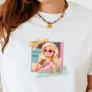 Puede incluir: Camiseta blanca con un gr&aacute;fico de una mujer rubia con gafas de sol rosas, sosteniendo una bebida. El texto "Caffeine" est&aacute; encima de la imagen, con "...is my personality" debajo. La camiseta la lleva una persona con vaqueros azules y una rebeca beige.
