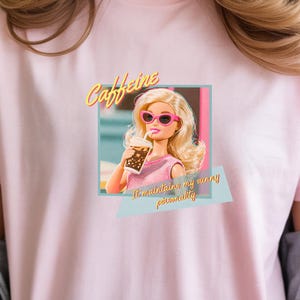 Puede incluir: Camiseta rosa claro con un estampado de una mu&ntilde;eca rubia con gafas de sol rosas y una bebida. El texto "Caffeine" est&aacute; encima de la mu&ntilde;eca, y "I'm maintaining my wing personality" est&aacute; debajo. Estilo casual.