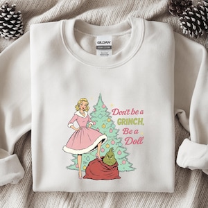 May include: Cream-colored sweatshirt with a graphic featuring a woman in a pink dress, a Christmas tree, and the Grinch. Text reads "Don't be a Grinch, Be a Doll". The design is in a cartoon style.