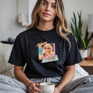 Puede incluir: Camiseta negra con un gr&aacute;fico de una mu&ntilde;eca con gafas de sol y sosteniendo una taza de caf&eacute;, con la palabra "Caffeine" arriba y texto abajo. La camiseta es de cuello redondo.