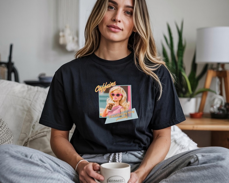Puede incluir: Camiseta negra con un gr&aacute;fico de una mu&ntilde;eca con gafas de sol y sosteniendo una taza de caf&eacute;, con la palabra "Caffeine" arriba y texto abajo. La camiseta es de cuello redondo.